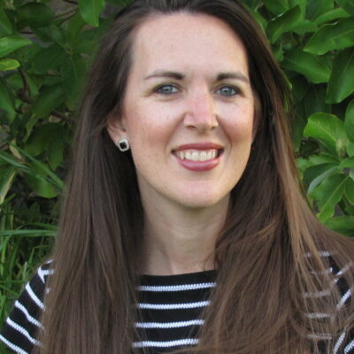 A woman in a striped shirt smiling in front of bushes.