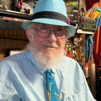 Elderly man with a white beard wearing a blue hat and turquoise shirt, smiling while seated indoors.