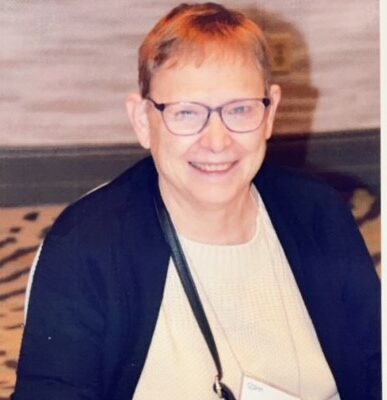 A person with short hair and glasses, wearing a name tag, sits at a table with a laptop, smiling at the camera.