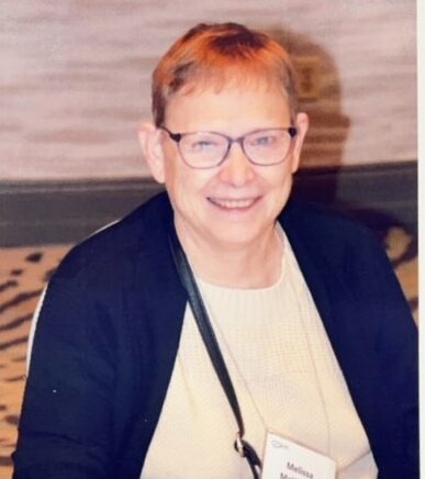 A person with short hair and glasses, wearing a name tag, sits at a table with a laptop, smiling at the camera.