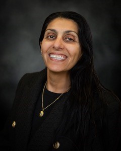 A person with long black hair smiles, wearing a black jacket and a necklace with a pendant, in front of a dark background.