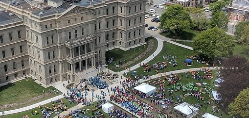 A large crowd gathers on the lawn of an ornate, multi-story building, with tents and tables visible. Trees and surrounding streets frame the scene.